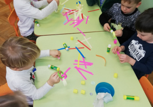 na zdjęciu dzieci siedzą przy stoliku, kleją łańcuch z papieru. Na stole materiały potrzebne do wykonania łańcucha: paski kolorowego papieru i kleje